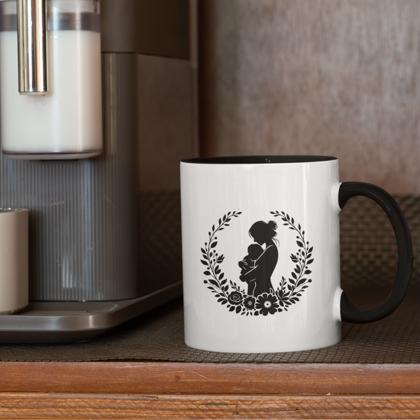 Beautiful white ceramic mug with a black interior and handle, showcasing a heartfelt silhouette design of a mother lovingly holding her baby, surrounded by a delicate floral wreath. This mug is perfect for celebrating motherhood. Ideal as a thoughtful gift for new moms, Mother’s Day, or baby showers. Microwave and dishwasher safe, this mug combines warmth, style, and durability for everyday use.