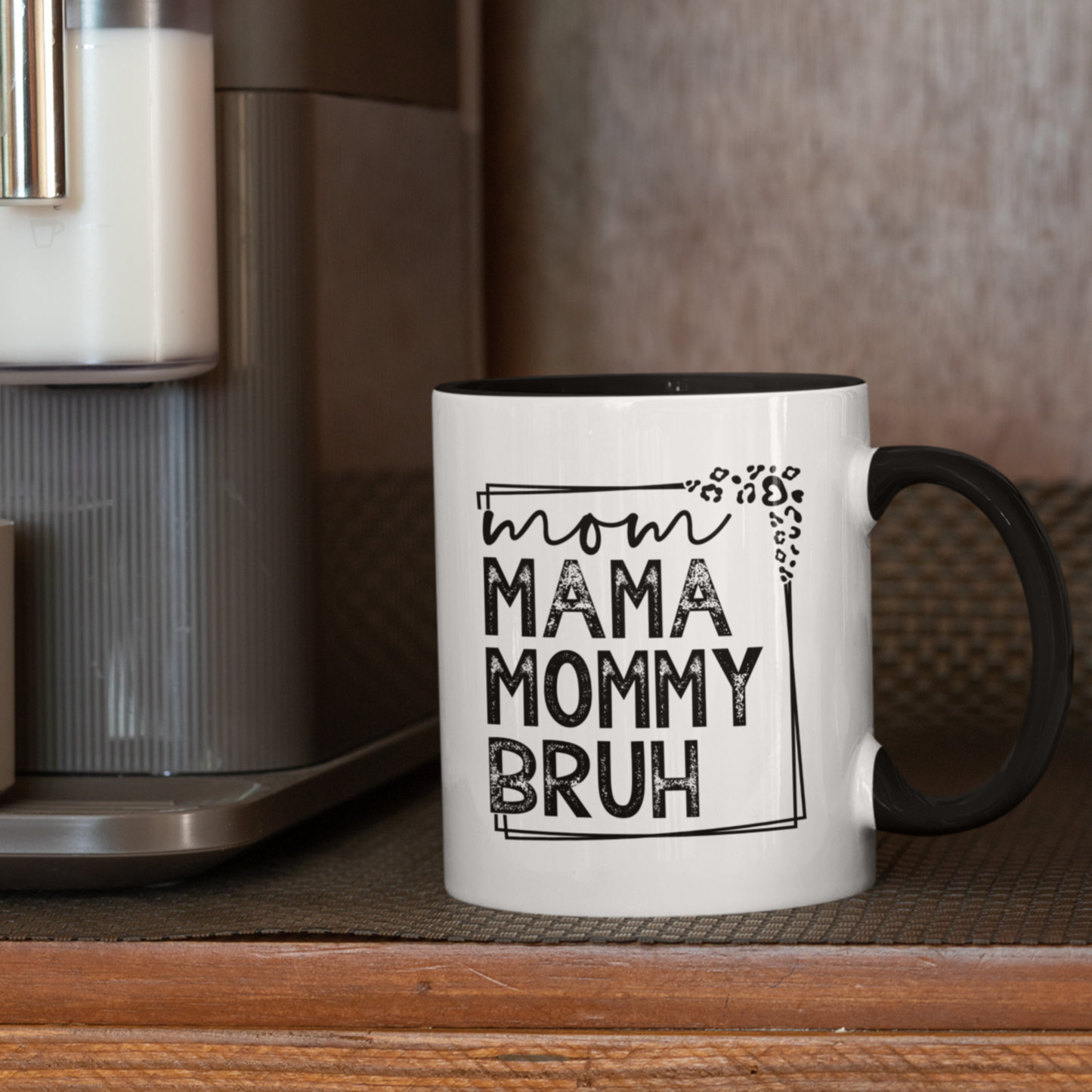 11 oz white ceramic mug with a black interior and handle. The mug features the black text "mom MAMA MOMMY BRUH" stacked vertically within a rectangular frame with leopard print accents. Dried citrus slices and small white flowers are arranged near the base of the mug.