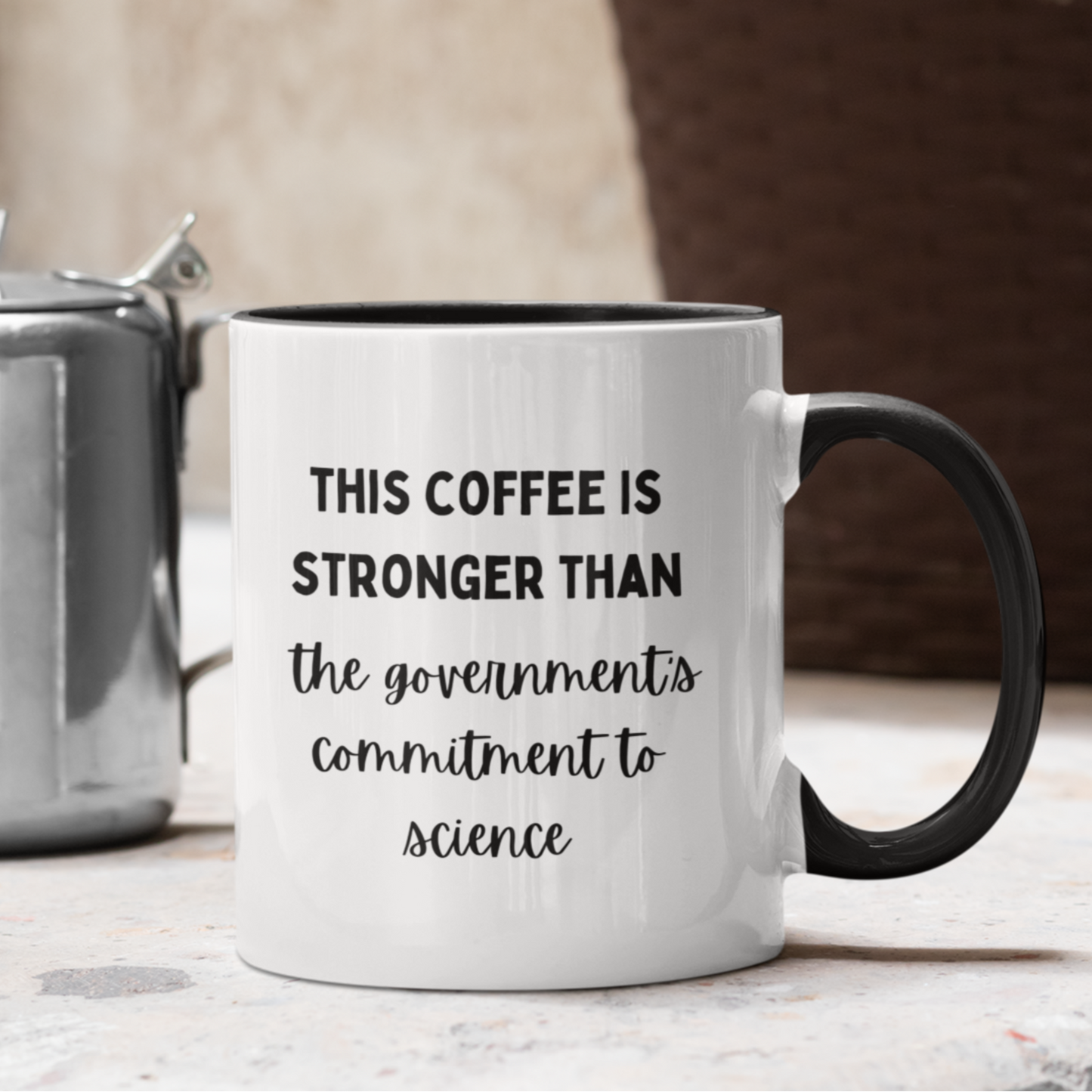11 oz white ceramic coffee mug with a glossy finish, featuring a sleek black handle and a matching black interior. The mug displays a humorous and bold printed message in a combination of fonts. The text reads: "THIS COFFEE IS STRONGER THAN the government's commitment to science." The top line is in bold uppercase, while the bottom lines are in a casual, handwritten-style script, adding a playful touch.