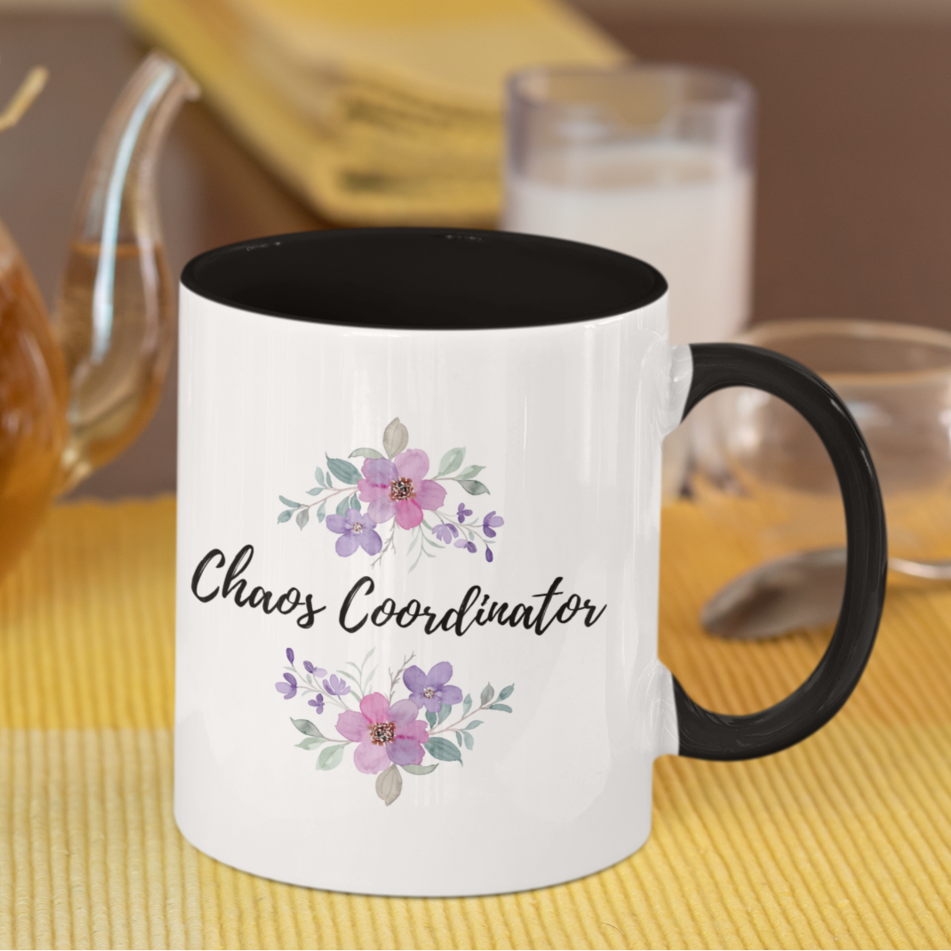 11 oz ceramic coffee mug featuring a sleek white exterior with a contrasting black interior and handle. The front design showcases elegant hand-painted floral illustrations in soft pastel shades of pink, purple, and lilac with delicate green leaves. Centered between the floral arrangements is the playful and stylish script text: “Chaos Coordinator.”