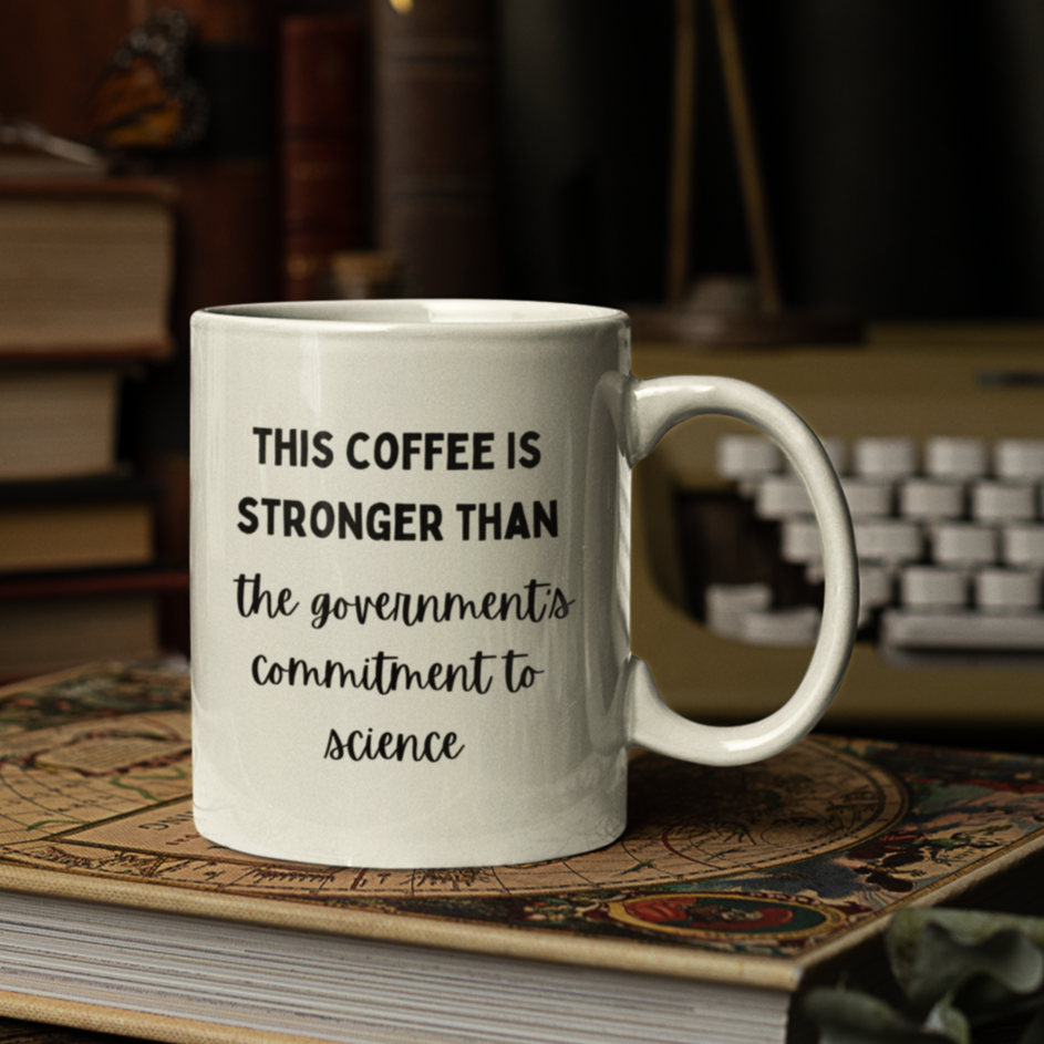 11 oz white ceramic coffee mug featuring a humorous and bold printed message in black typography. The text reads, "THIS COFFEE IS STRONGER THAN the government's commitment to science," with varying font styles—bold uppercase for emphasis and cursive script for a playful contrast.