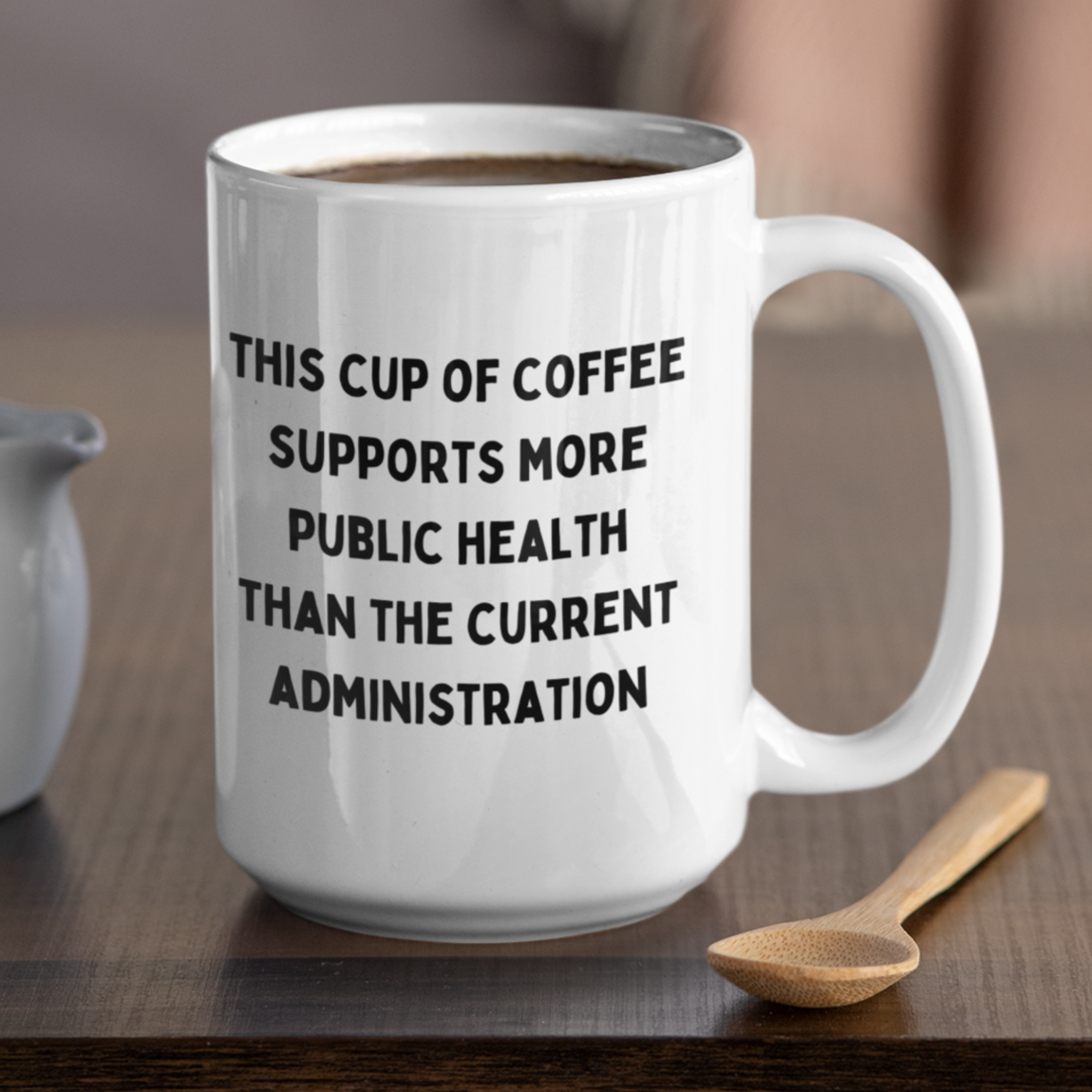 15 oz white ceramic coffee mug with handle, perfect for daily use at home or in the office. The mug features a bold, humorous political statement printed in all-caps black font: "THIS CUP OF COFFEE SUPPORTS MORE PUBLIC HEALTH THAN THE CURRENT ADMINISTRATION." The text is centered and covers most of the front surface, making it highly visible.