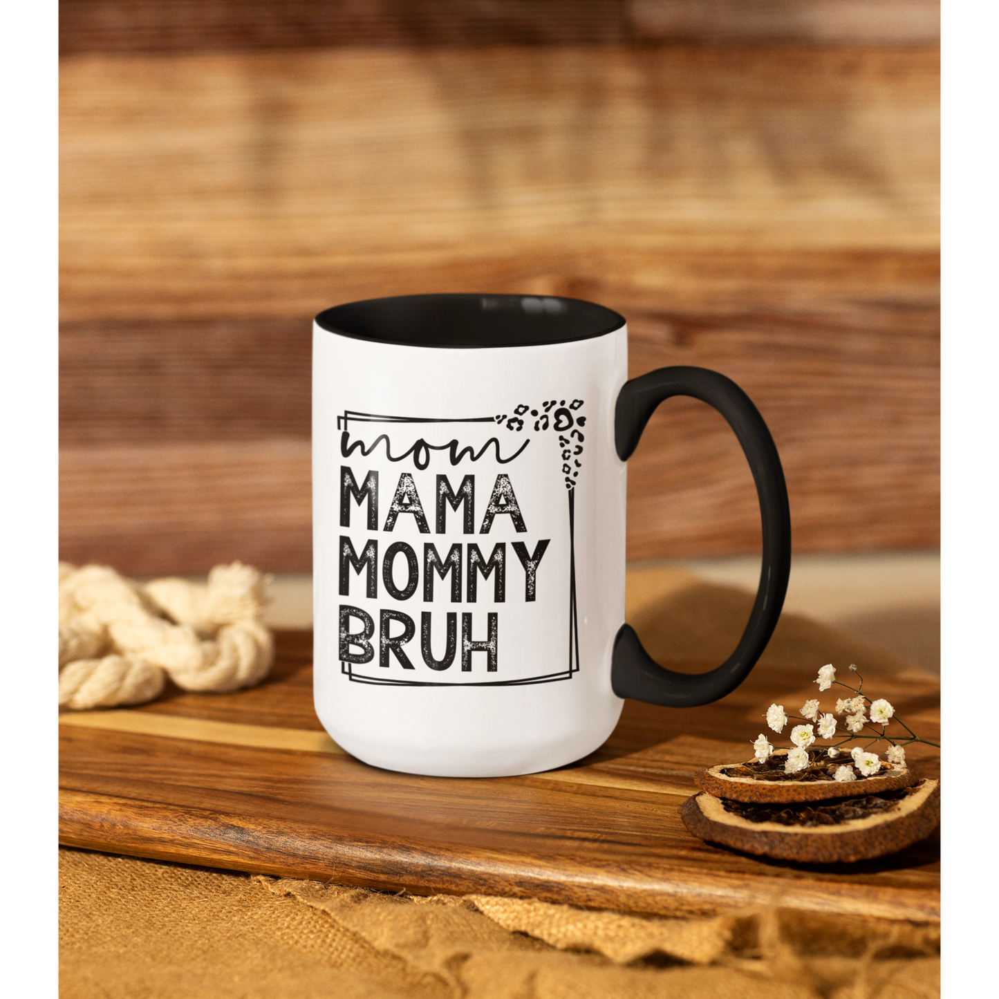 15 oz white ceramic mug with a black interior and handle. The mug features the black text "mom MAMA MOMMY BRUH" stacked vertically within a rectangular frame with leopard print accents. Dried citrus slices and small white flowers are arranged near the base of the mug.