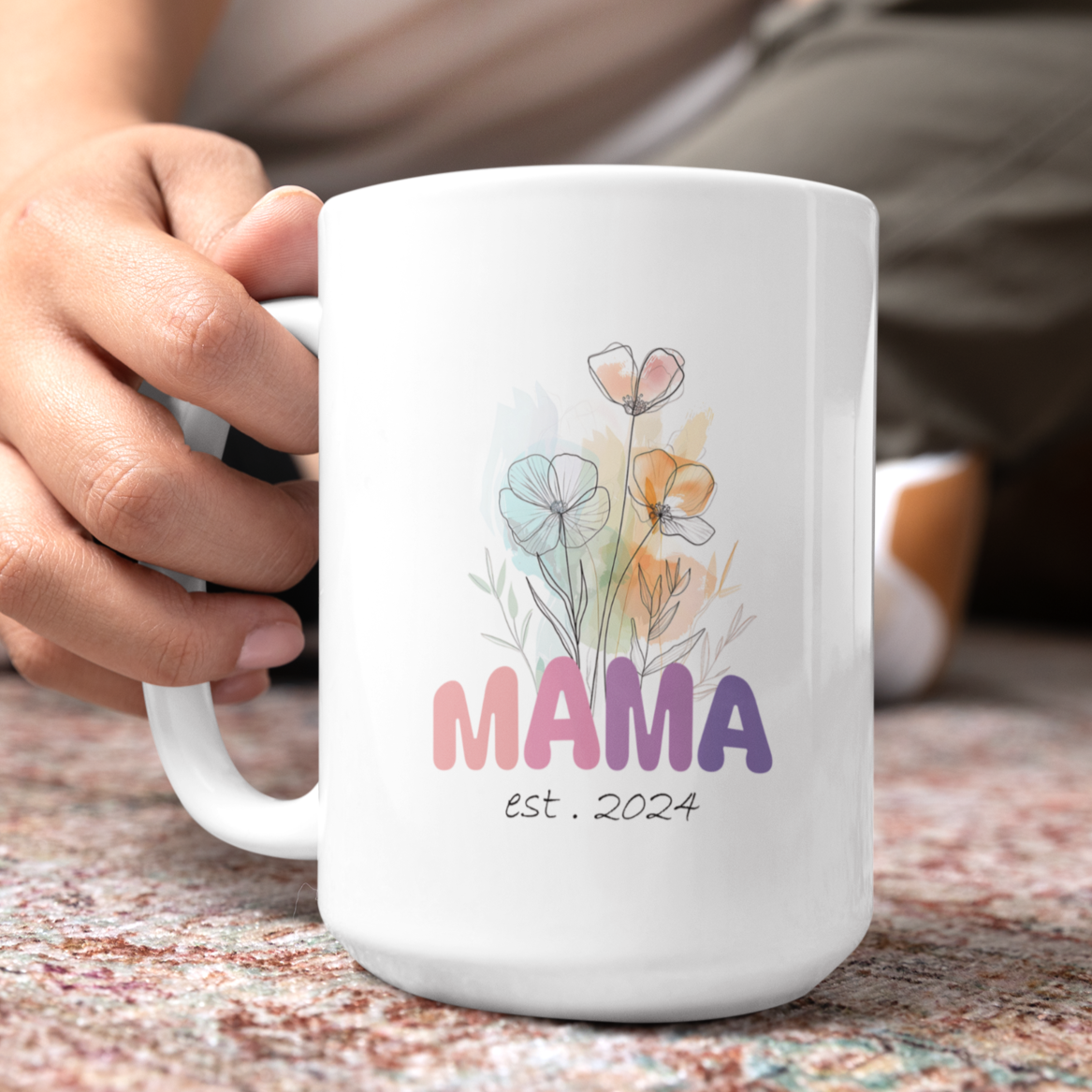 Charming 15 oz white ceramic mug featuring a soft watercolor-style floral bouquet and the word “MAMA” in bold, colorful letters transitioning from pink to purple. Beneath the floral artwork, the text reads “est. 2024” in a delicate handwritten font. Perfect gift for new moms or a Mother’s Day surprise celebrating motherhood’s newest chapter.