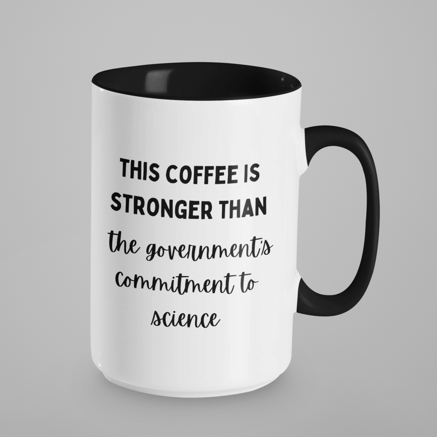 15 oz white ceramic coffee mug with a glossy finish, featuring a sleek black handle and a matching black interior. The mug displays a humorous and bold printed message in a combination of fonts. The text reads: "THIS COFFEE IS STRONGER THAN the government's commitment to science." The top line is in bold uppercase, while the bottom lines are in a casual, handwritten-style script, adding a playful touch.