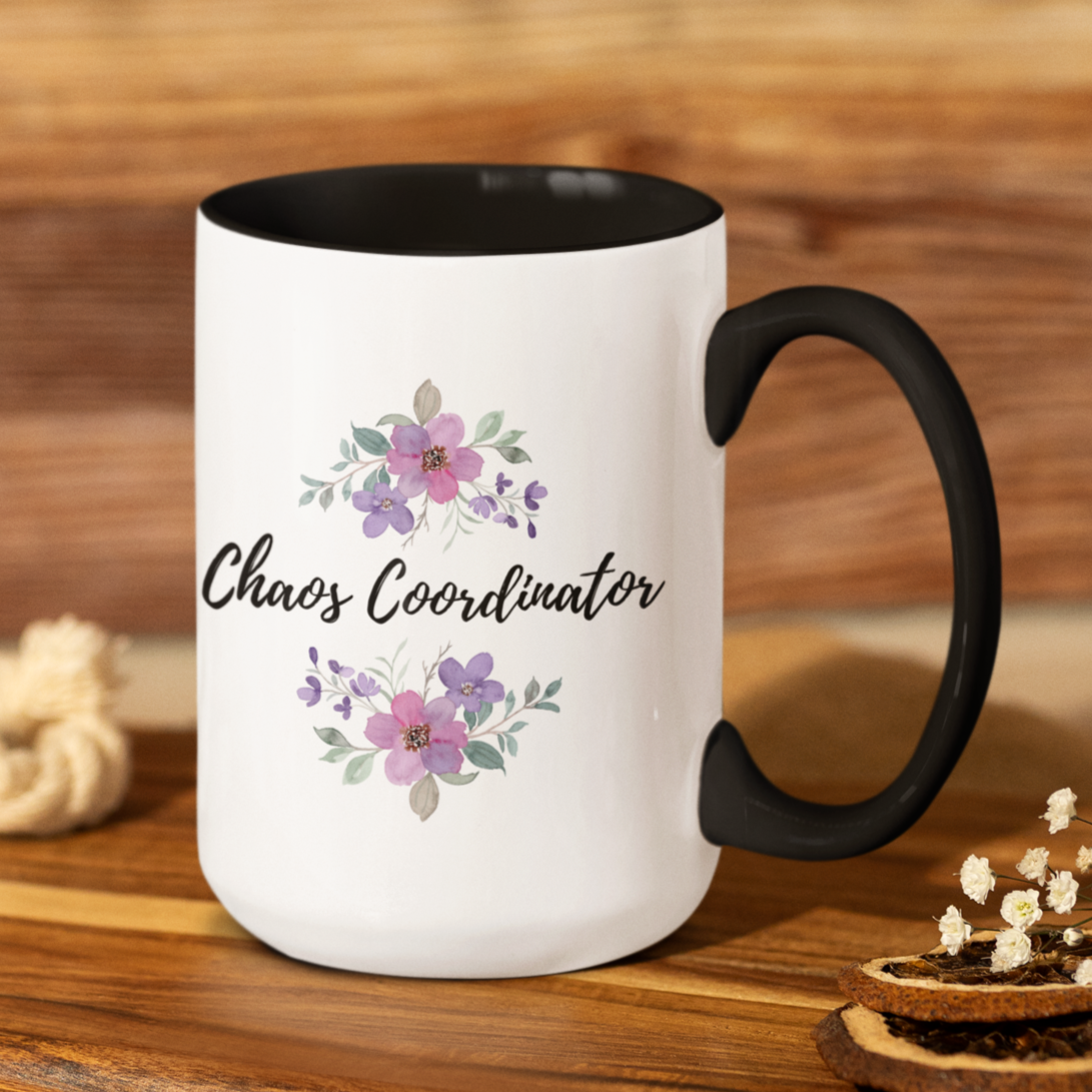 15 oz ceramic coffee mug featuring a sleek white exterior with a contrasting black interior and handle. The front design showcases elegant hand-painted floral illustrations in soft pastel shades of pink, purple, and lilac with delicate green leaves. Centered between the floral arrangements is the playful and stylish script text: “Chaos Coordinator.”