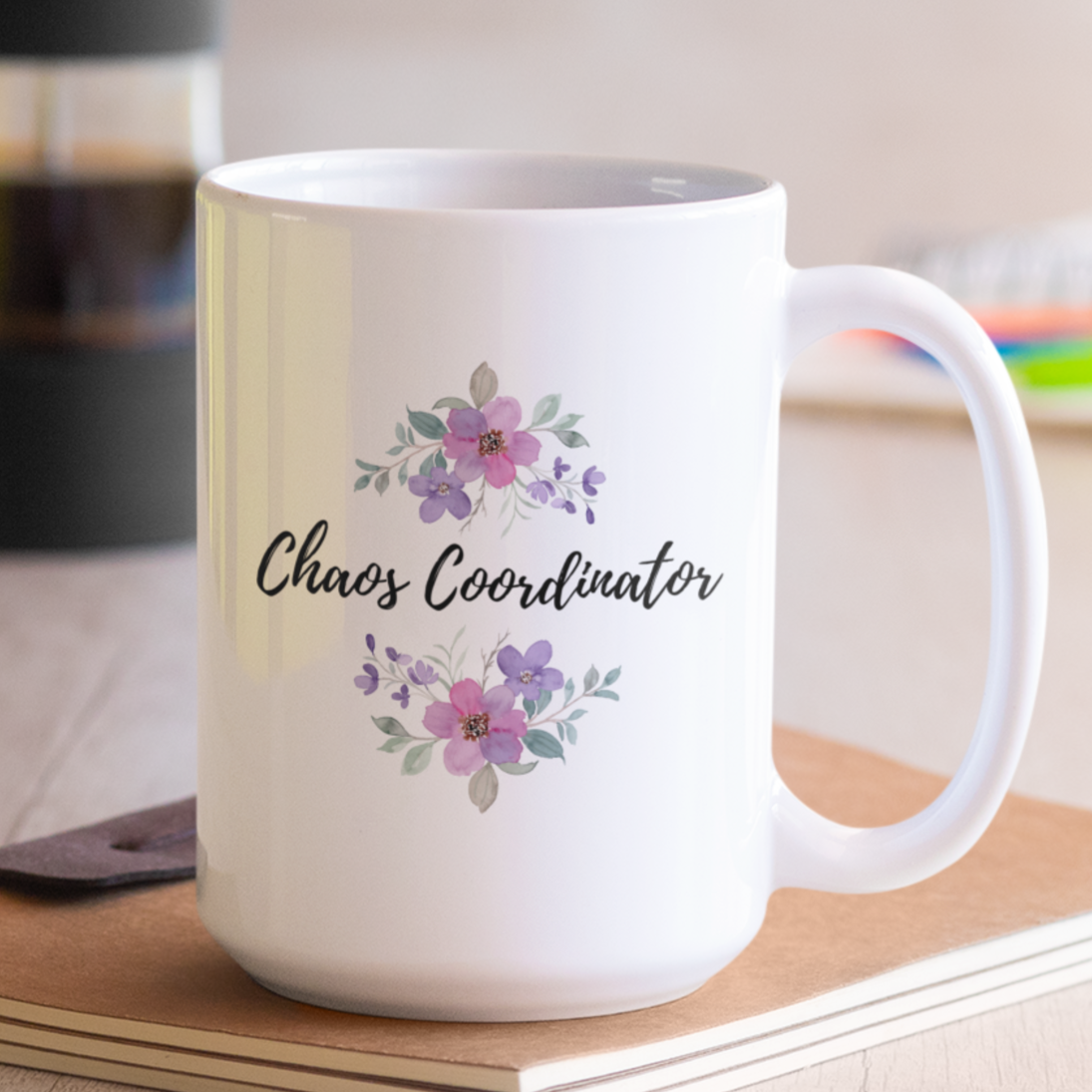 15 oz white ceramic coffee mug featuring the playful phrase “Chaos Coordinator” in stylish black script font centered on the front. Surrounding the text are delicate watercolor floral arrangements in shades of soft pink, lavender, and muted green leaves, adding a touch of elegance and charm. The mug has a smooth glossy finish and a sturdy C-shaped handle for easy grip.