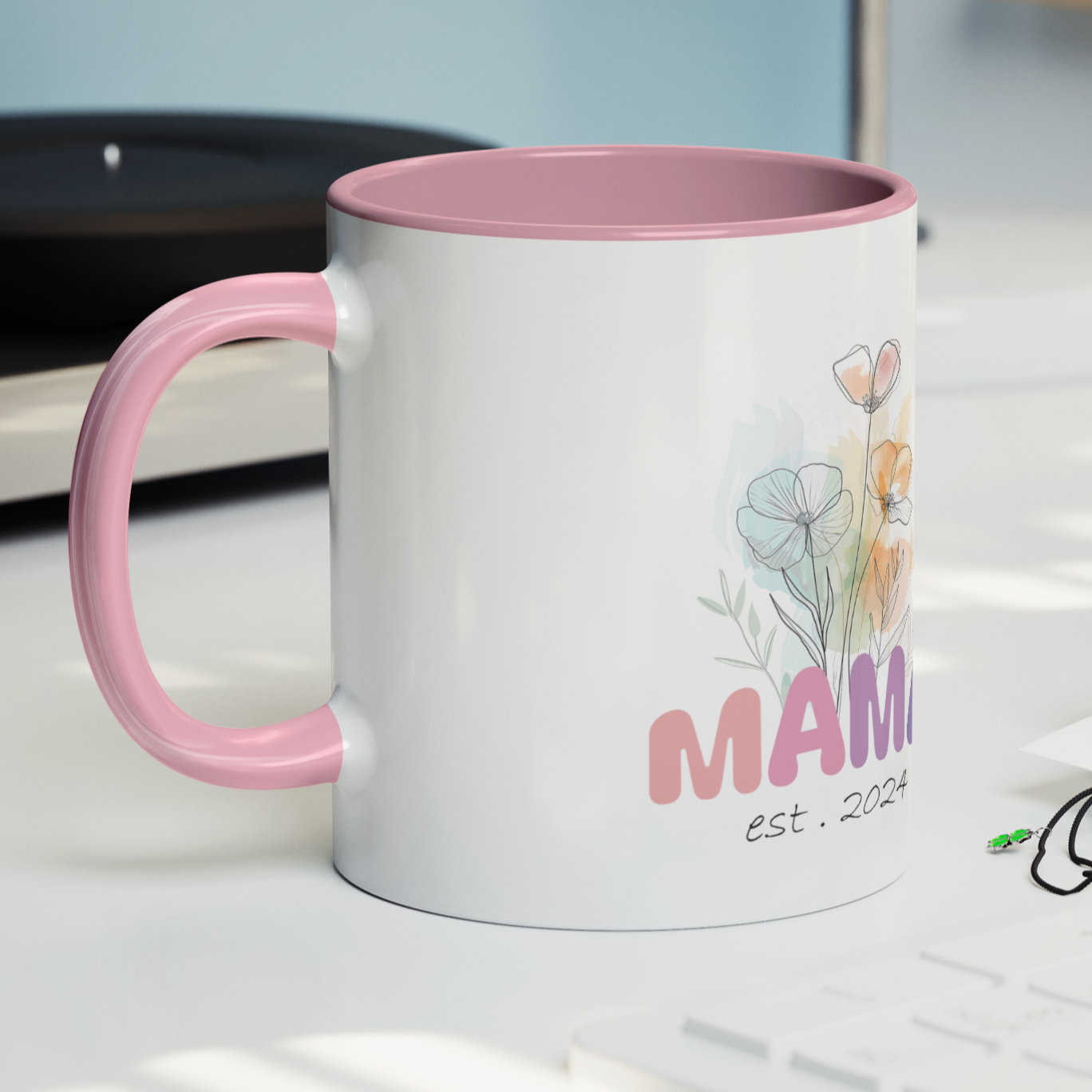 Adorable 11 oz white ceramic coffee mug with a glossy finish, featuring a soft pink interior and matching pink handle. The design showcases a delicate watercolor floral bouquet above the word “MAMA” in cheerful gradient tones from pink to purple, with the text “est. 2024” in a stylish handwritten script below. This mug is a perfect gift for new moms celebrating their first Mother's Day or a sweet keepsake marking the beginning of their motherhood journey.