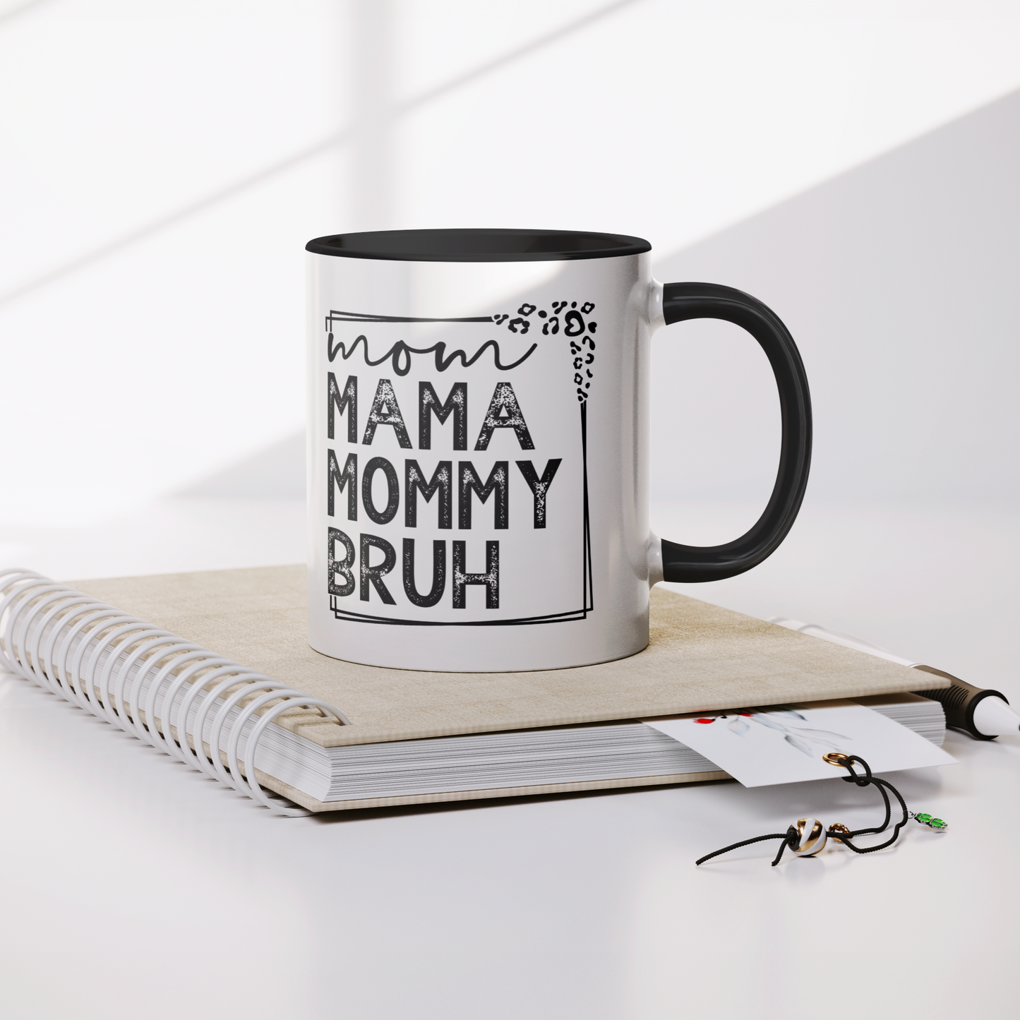 11 oz white ceramic mug with a black interior and handle. The mug features the black text "mom MAMA MOMMY BRUH" stacked vertically within a rectangular frame with leopard print accents. Dried citrus slices and small white flowers are arranged near the base of the mug.