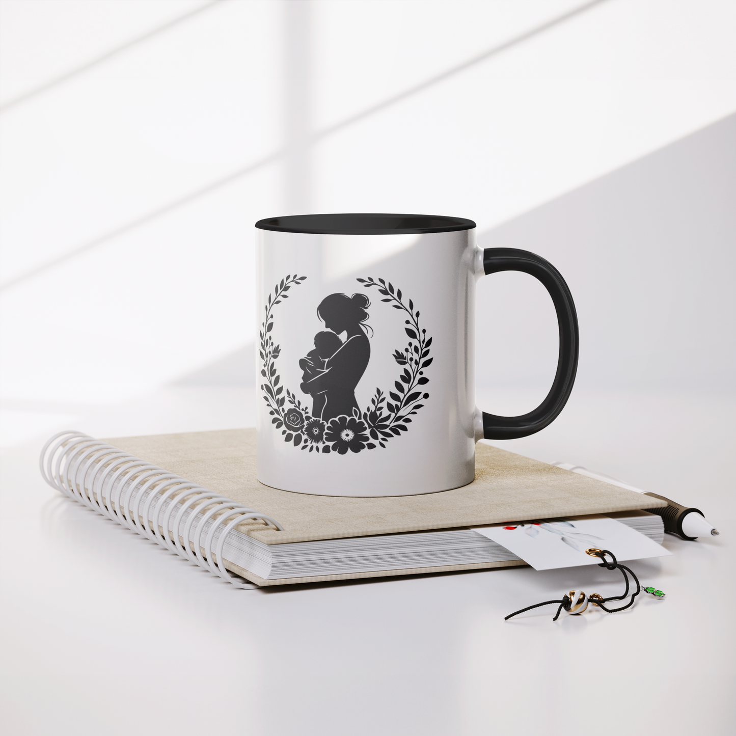 Beautiful white ceramic mug with a black interior and handle, showcasing a heartfelt silhouette design of a mother lovingly holding her baby, surrounded by a delicate floral wreath. This mug is perfect for celebrating motherhood. Ideal as a thoughtful gift for new moms, Mother’s Day, or baby showers. Microwave and dishwasher safe, this mug combines warmth, style, and durability for everyday use.