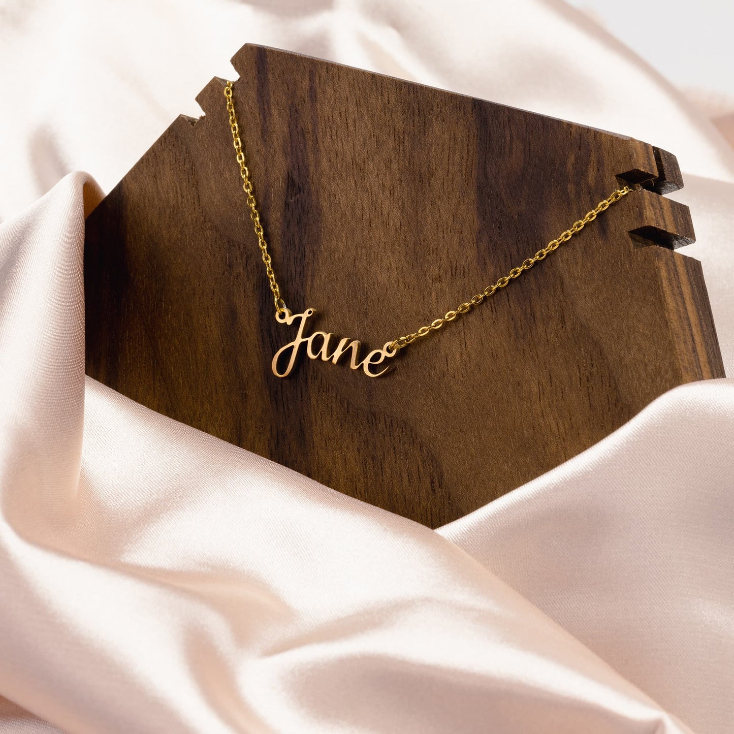 A luxurious, personalized gold name necklace featuring the name "Jane" in elegant cursive script. The necklace is showcased on a dark wooden display stand with a smooth, rich grain that enhances the necklace's golden shine. The setup is complemented by soft, cream-colored satin fabric, creating an elegant and sophisticated backdrop. Perfect as a custom gift for birthdays, anniversaries, or special occasions.