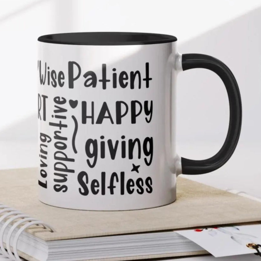 Elegant white ceramic mug with a black handle and black interior, featuring a bold, black hand-lettered word cloud celebrating motherhood. Words like “Wise,” “Patient,” “Happy,” “Giving,” “Selfless,” “Supportive,” and “Loving” wrap around the mug in playful, artistic fonts. The mug is staged on a spiral notebook with a pen and beaded charm, evoking a cozy, thoughtful vibe. Perfect gift for Mother’s Day, birthdays, or to honor the strength and love of any mom.