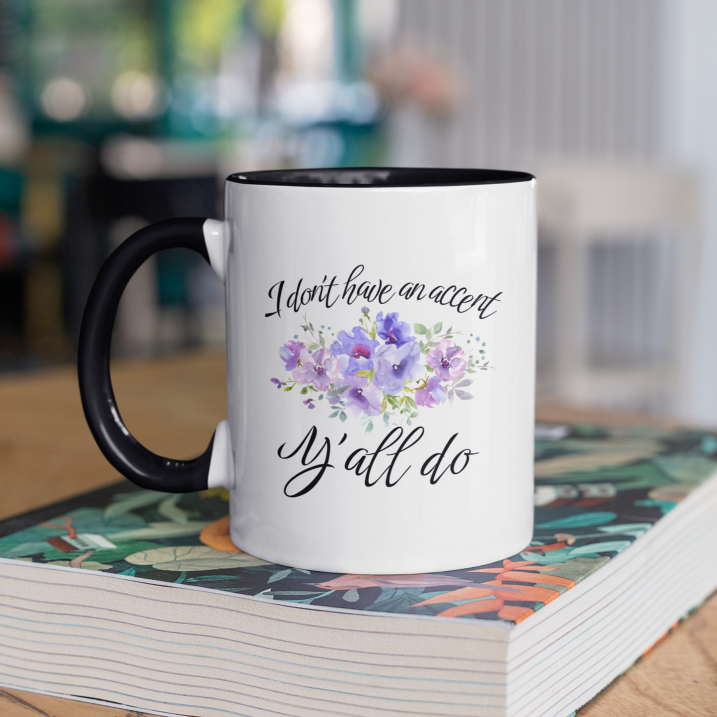 11 oz stylish ceramic coffee mug featuring a white glossy exterior with a contrasting black handle and black interior. The front of the mug displays elegant, cursive black text that reads, "I don't have an accent, y'all do," accented by a beautiful watercolor floral arrangement in shades of purple, lilac, and soft green.