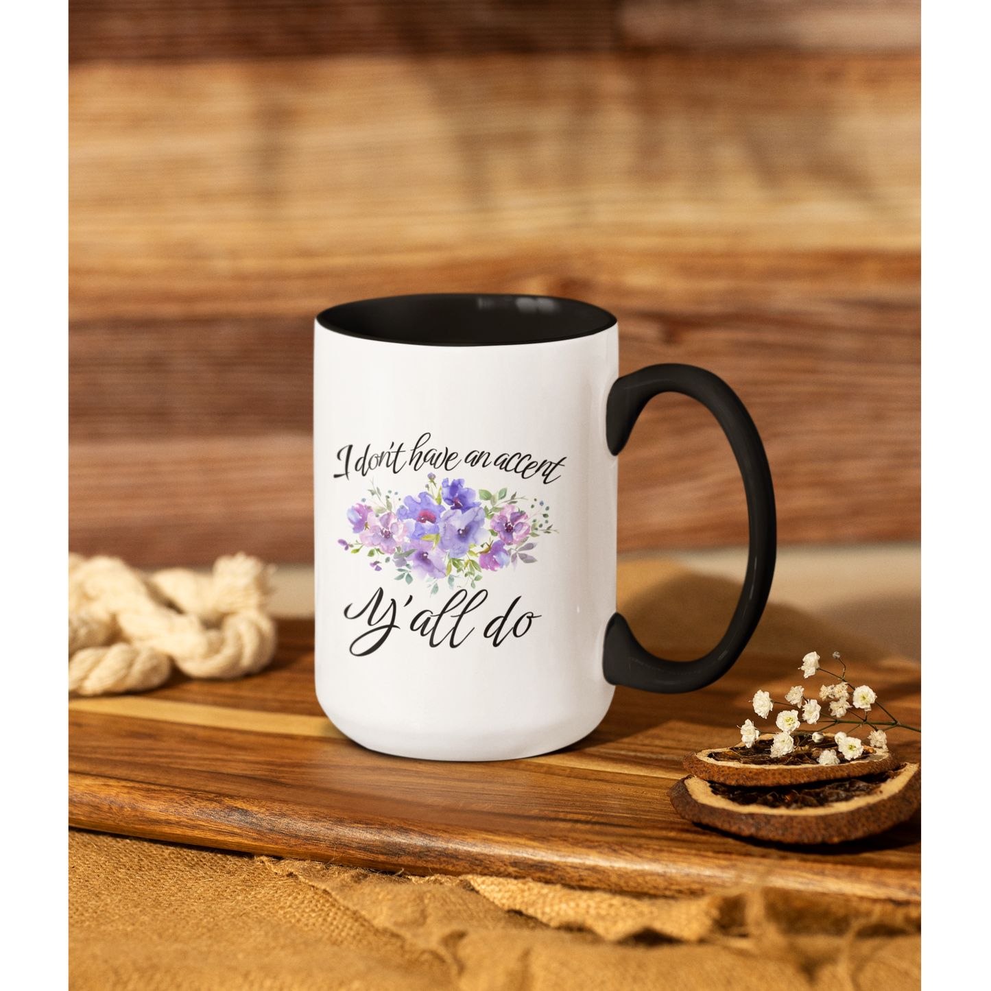 15 oz stylish ceramic coffee mug featuring a white glossy exterior with a contrasting black handle and black interior. The front of the mug displays elegant, cursive black text that reads, "I don't have an accent, y'all do," accented by a beautiful watercolor floral arrangement in shades of purple, lilac, and soft green.