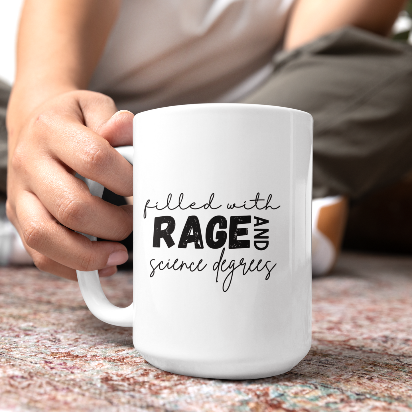 15 oz Close-up of a white ceramic coffee mug that features a bold and playful typography design that reads: "filled with RAGE AND science degrees." The words "RAGE AND" are in bold, uppercase letters, while the rest of the text is in a casual, handwritten script. Ideal for scientists, academics, or STEM enthusiasts.