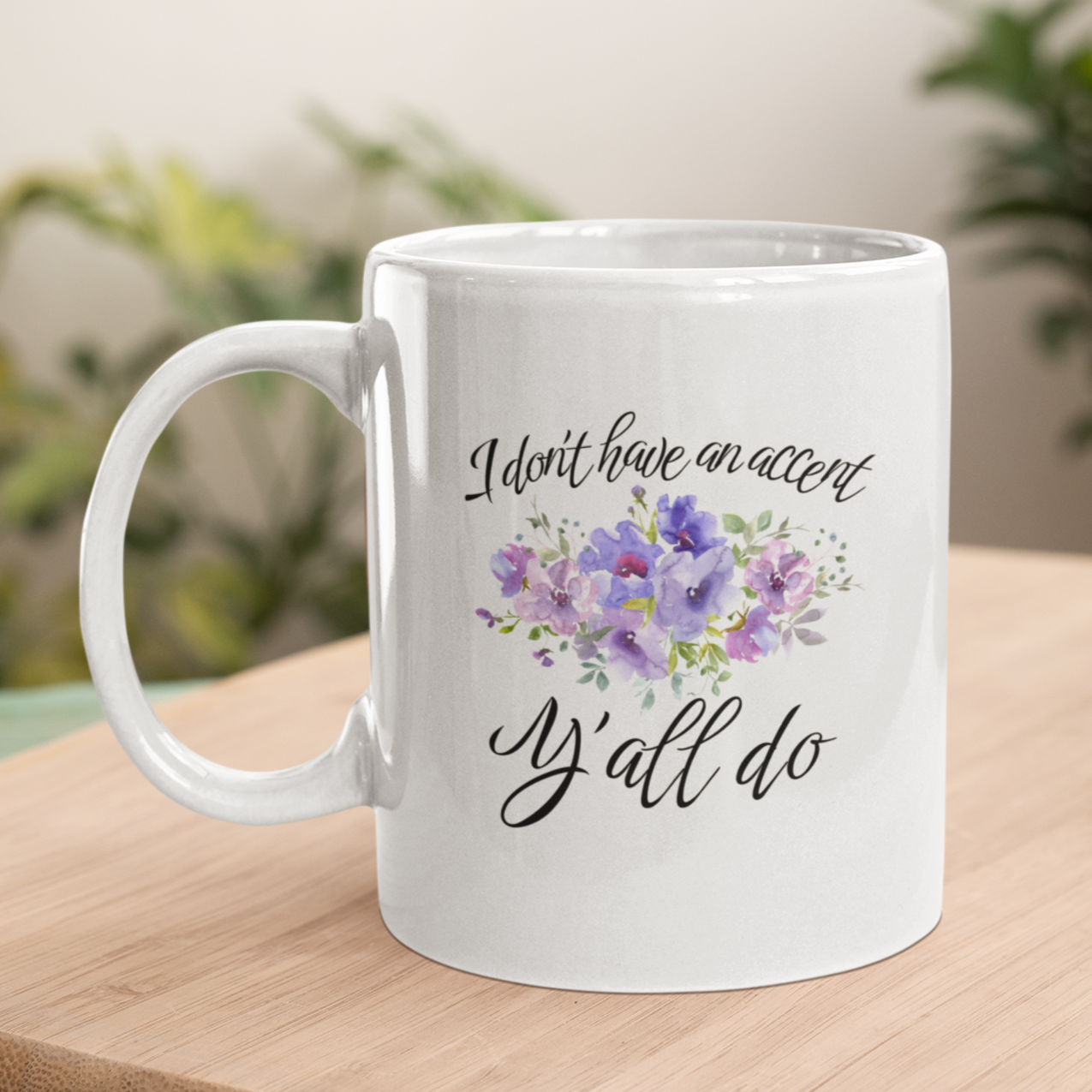 11 oz White ceramic coffee mug featuring a charming, southern-inspired design. The front showcases a whimsical quote in elegant, black cursive font that reads, “I don’t have an accent, Y’all do.” Between the two lines of text, there’s a delicate watercolor illustration of purple and lavender flowers with green foliage, adding a soft, feminine touch.
