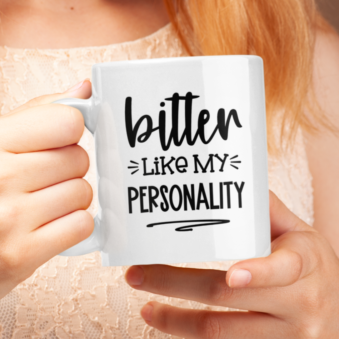 Humorous 11 oz white ceramic mug held in two hands. Black text reads "bitter LIKE MY PERSONALITY" in a playful, slightly sarcastic font. Perfect for those with a dry wit and a love for coffee or tea. Makes a funny gift for friends, family, or coworkers who appreciate dark humor.