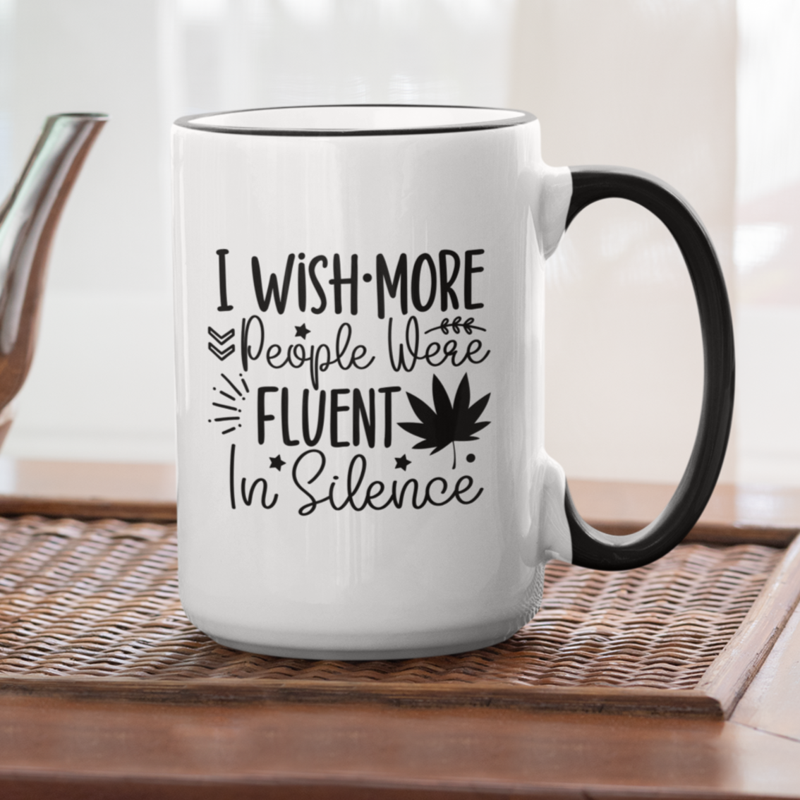 Charming white ceramic mug with a black interior and matching handle, featuring a witty black typography design that says, “I wish more people were fluent in silence,” alongside playful icons like stars, leaves, and a bold maple leaf silhouette. A perfect gift for introverts, tea lovers, or anyone who appreciates peace and clever design.