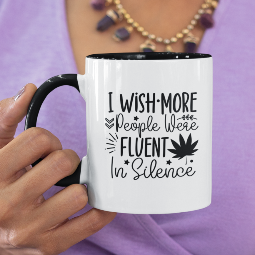 Charming white ceramic mug with a black interior and matching handle, featuring a witty black typography design that says, “I wish more people were fluent in silence,” alongside playful icons like stars, leaves, and a bold maple leaf silhouette. A perfect gift for introverts, tea lovers, or anyone who appreciates peace and clever design.