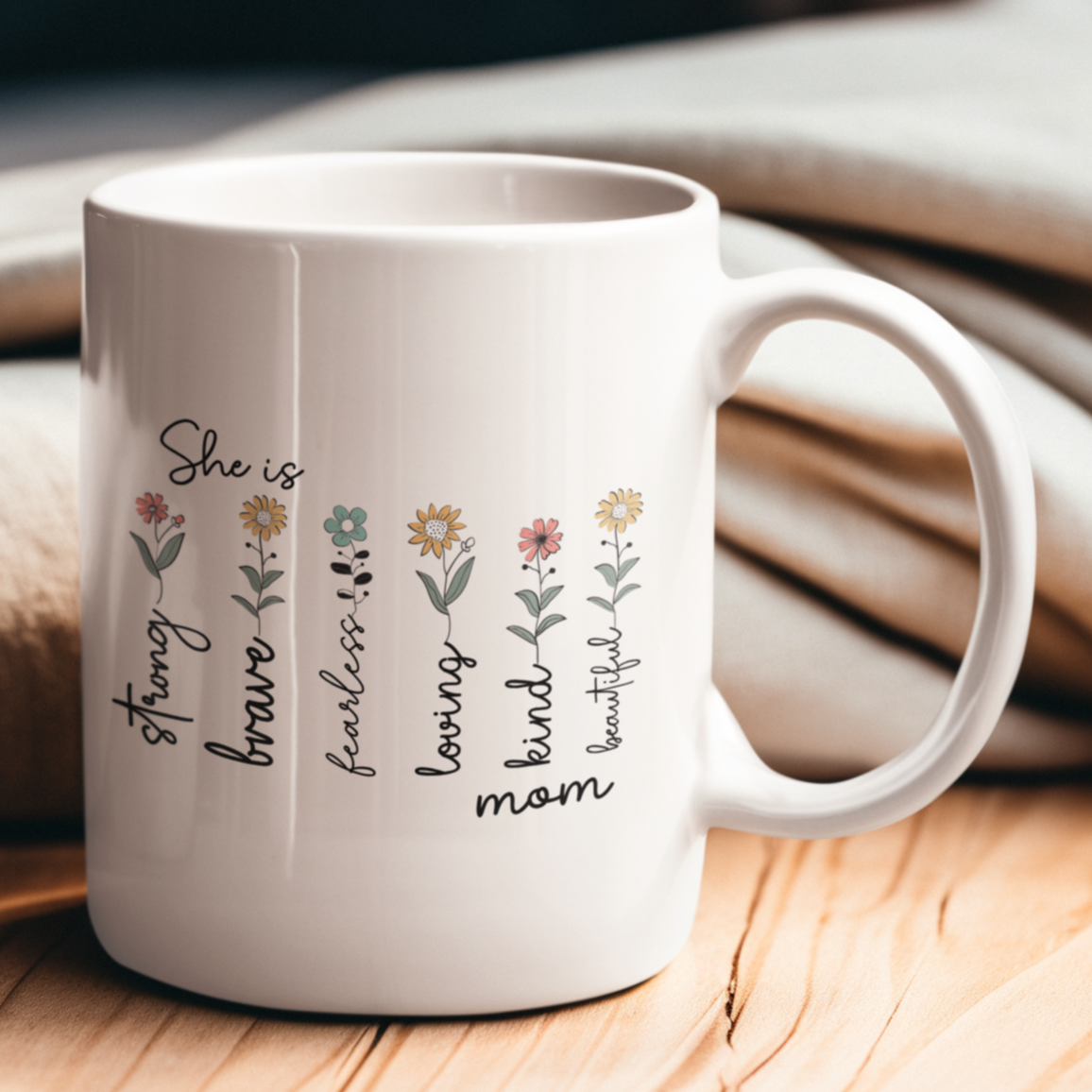 Elegant white ceramic coffee mug featuring a heartwarming floral design with the words “She is strong, brave, fearless, loving, kind, mom, beautiful” written alongside delicate illustrated flowers. The mug has a classic shape with a large, comfortable handle, making it both functional and beautiful. Displayed on top of stacked books with a pair of reading glasses nearby, the setting evokes a cozy, thoughtful vibe — perfect for gifting to moms, grandmothers, or inspirational women.