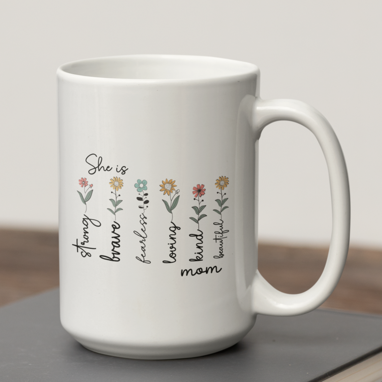 Elegant white ceramic coffee mug featuring a heartwarming floral design with the words “She is strong, brave, fearless, loving, kind, mom, beautiful” written alongside delicate illustrated flowers. The mug has a classic shape with a large, comfortable handle, making it both functional and beautiful. Displayed on top of stacked books with a pair of reading glasses nearby, the setting evokes a cozy, thoughtful vibe — perfect for gifting to moms, grandmothers, or inspirational women.