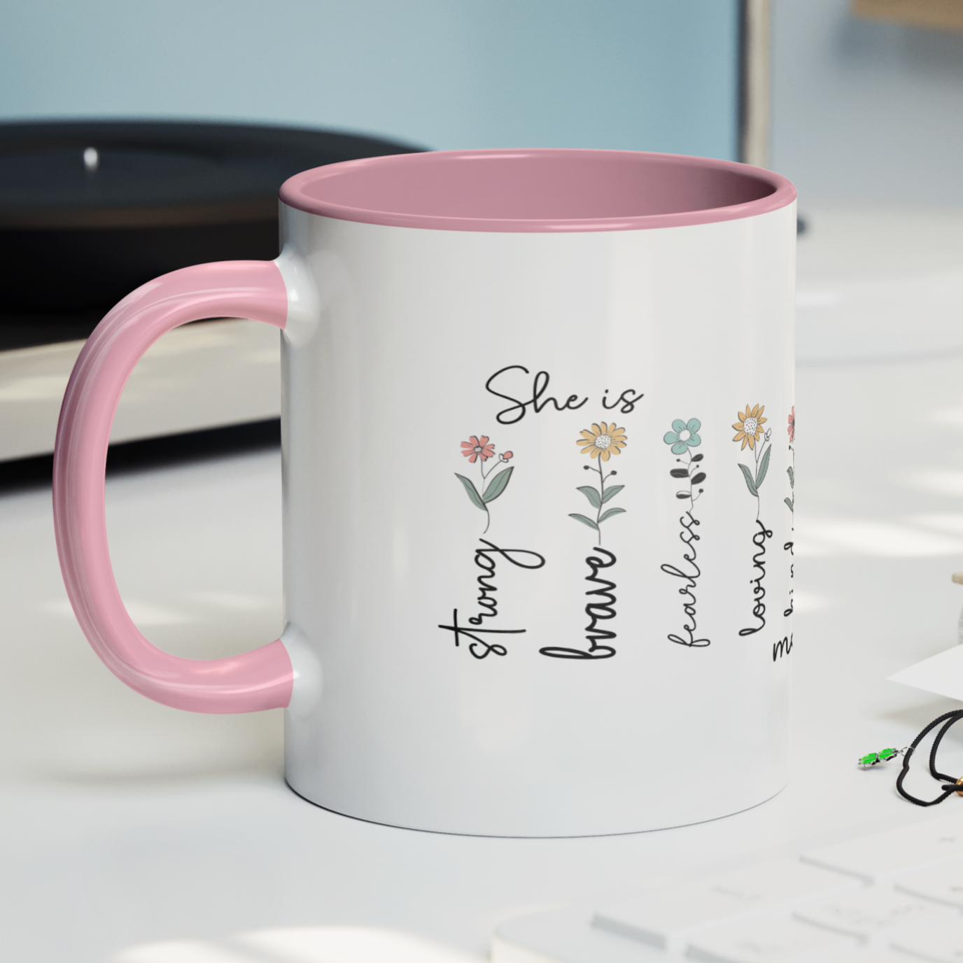 Charming 11 oz ceramic coffee mug with a glossy white exterior, soft pink interior, and matching pink handle. The design features delicate hand-drawn flowers paired with uplifting words that read: “She is strong, brave, fearless, loving, kind, mom, beautiful.” The heartfelt text and pastel floral accents create a warm, inspirational gift perfect for Mother's Day, birthdays, or everyday appreciation.