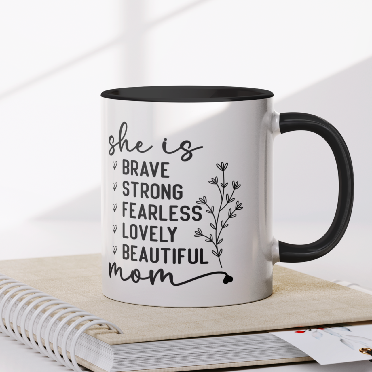 11 oz white ceramic mug with a black interior and handle. The mug features the black text 'she is BRAVE STRONG FEARLESS LOVELY BEAUTIFUL mom' stacked vertically in a handwritten-style font, accompanied by a delicate floral illustration.
