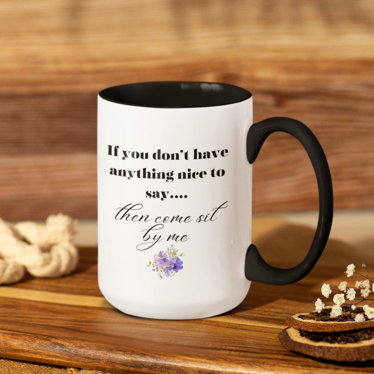 15 oz White ceramic coffee mug with a black handle and interior, featuring a playful and humorous quote printed in elegant black fonts. The text reads: "If you don’t have anything nice to say... then come sit by me." The phrase is styled with a combination of bold serif and flowing script fonts for emphasis. Below the quote, there’s a charming floral graphic with soft purple and pink flowers accented by green leaves.
