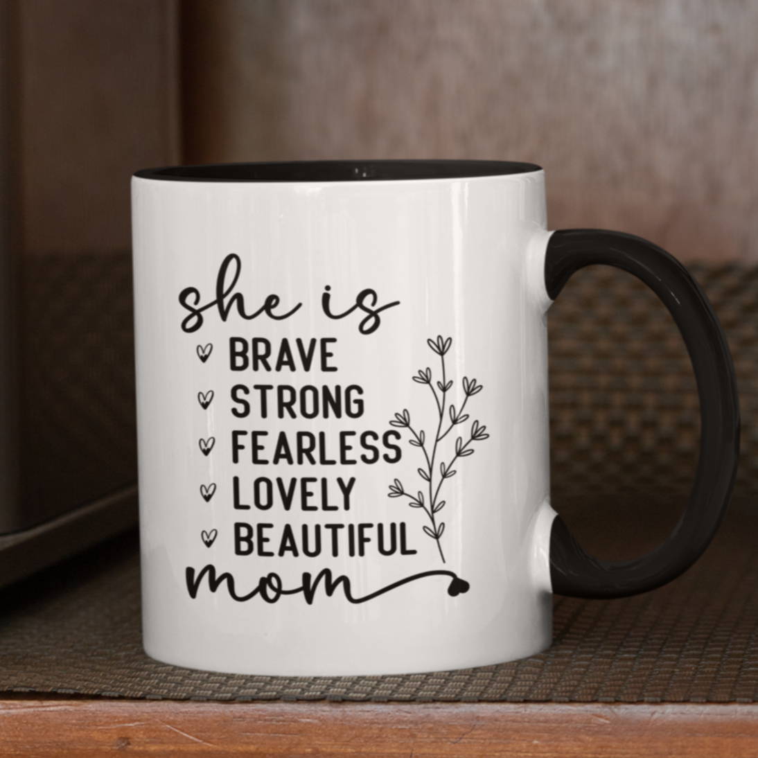 11 oz white ceramic mug with a black interior and handle. The mug features the black text 'she is BRAVE STRONG FEARLESS LOVELY BEAUTIFUL mom' stacked vertically in a handwritten-style font, accompanied by a delicate floral illustration.