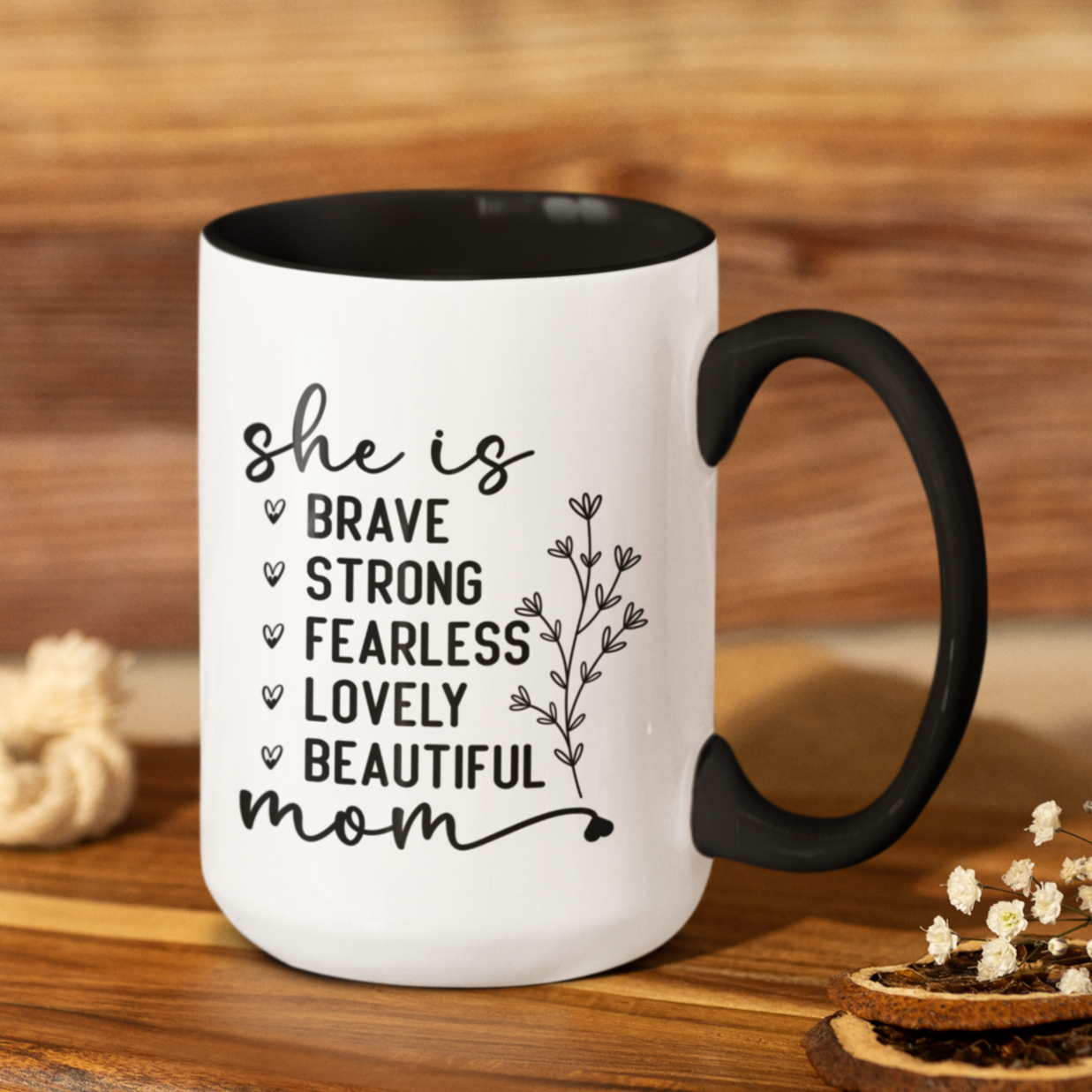 15 oz white ceramic mug with a black interior and handle. The mug features the black text 'she is BRAVE STRONG FEARLESS LOVELY BEAUTIFUL mom' stacked vertically in a handwritten-style font, accompanied by a delicate floral illustration.